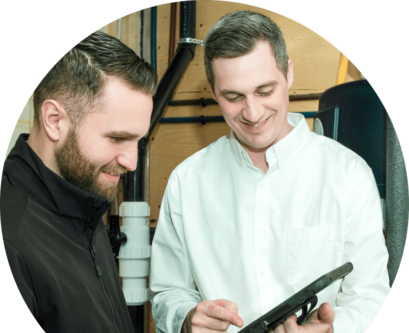 The Comfort Group specialist reviewing maintenance plan options with a smiling homeowner using a tablet, inside a utility room with visible piping. The Comfort Group specialist reviewing maintenance plan options with a smiling homeowner using a tablet, inside a utility room with visible piping.