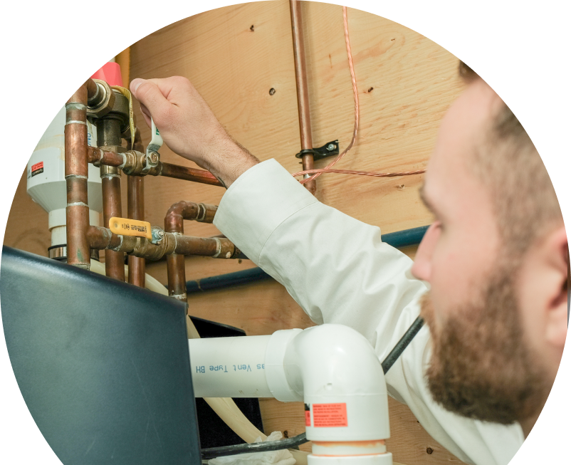 The Comfort Group water heater specialist tightens a valve on copper piping during a water heater installation, with exposed wood paneling and wiring in the background. The Comfort Group water heater specialist tightens a valve on copper piping during a water heater installation, with exposed wood paneling and wiring in the background.