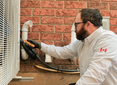 A The Comfort Group HVAC specialist wearing glasses and gloves uses a diagnostic tool to service an outdoor AC unit beside a brick wall. A The Comfort Group HVAC specialist wearing glasses and gloves uses a diagnostic tool to service an outdoor AC unit beside a brick wall.