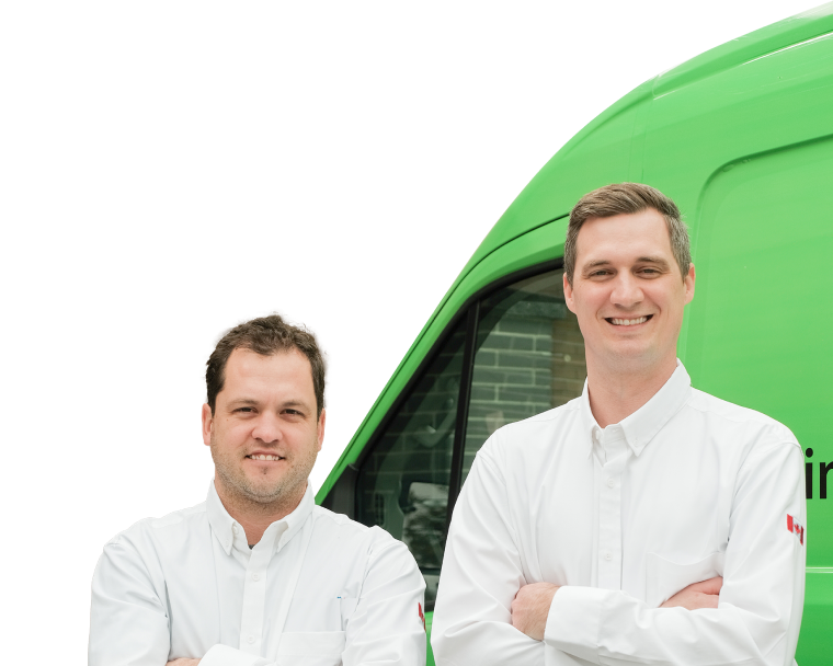 Two The Comfort Group technicians smiling in front of their green service van, wearing issued jackets during an on-site consultation.
