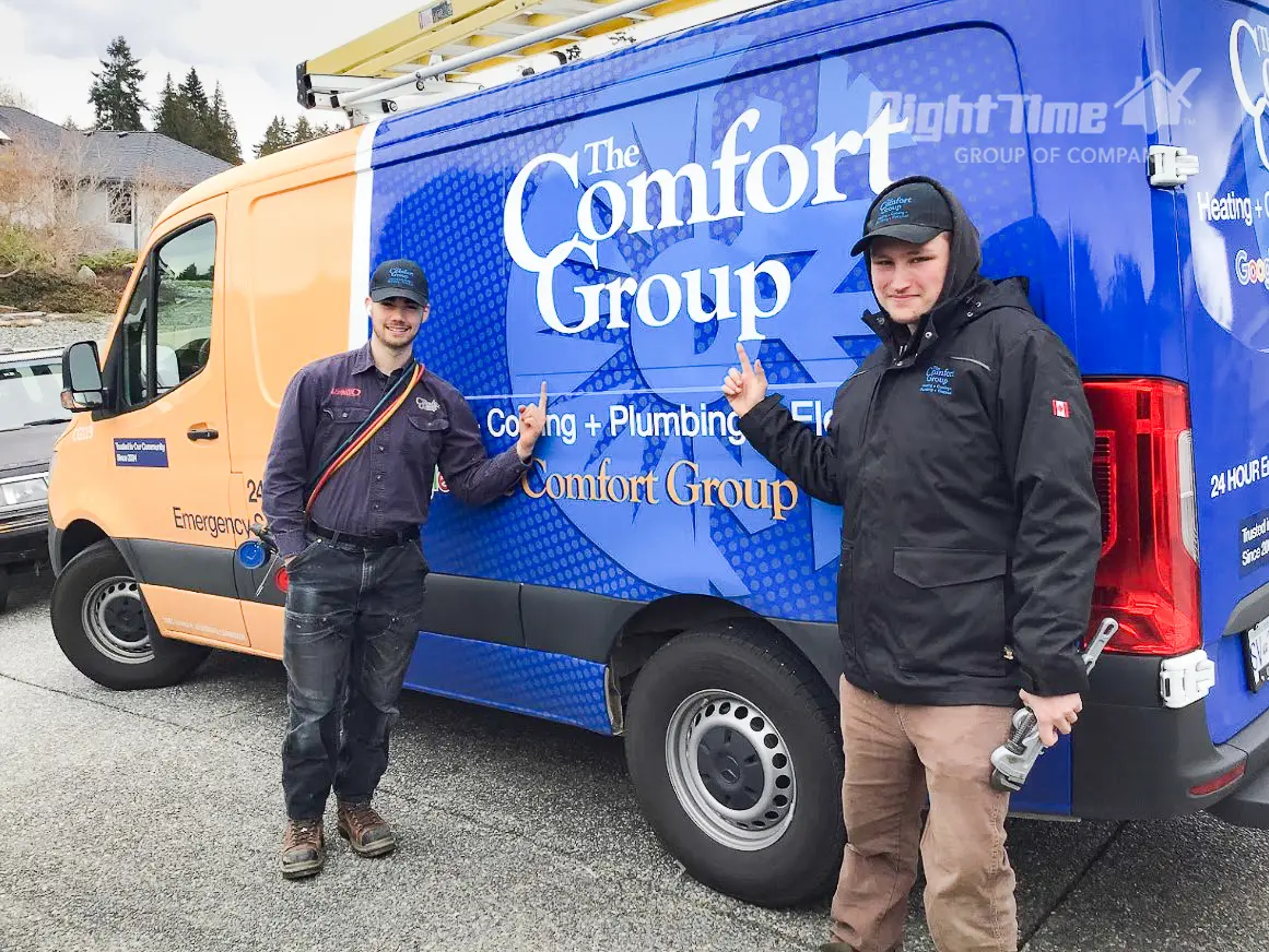Two of The Comfort Group technicians are standing by a branded service van on a rainy day, pointing to the logo on a residential street.
